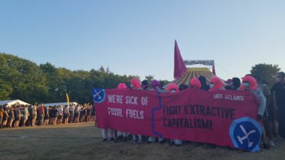 Eine lange Reihe von Menschen hinter einen transparent, im Hintergrund ein Zirkuszelt. Auf dem Transpi steht: We're sich of dossil fuels, fight extractive capitalism"