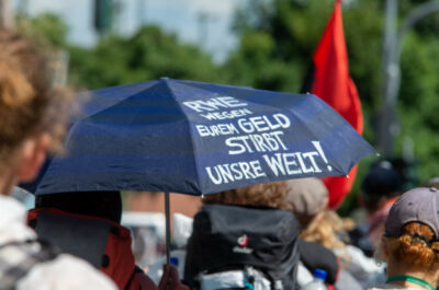 Aktivist*innen sind auf einer Demo unterwegs, ein Mensch hält einen Schirm auf dem steht "RWE wegen eurem Geld stirbt die Welt!