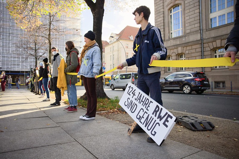 Aktivist*innen spannen ein gelbes Band um Lützerath, auf einem Schild steht "124 Jahre RWE sind genug"